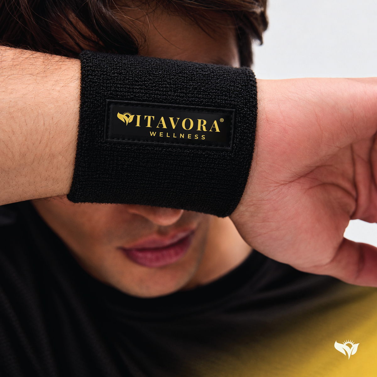 Close-up lifestyle shot of a Vitavora Wellness Power Circle Wristband worn on the wrist, featuring the gold embroidered logo patch against black terry cloth fabric.